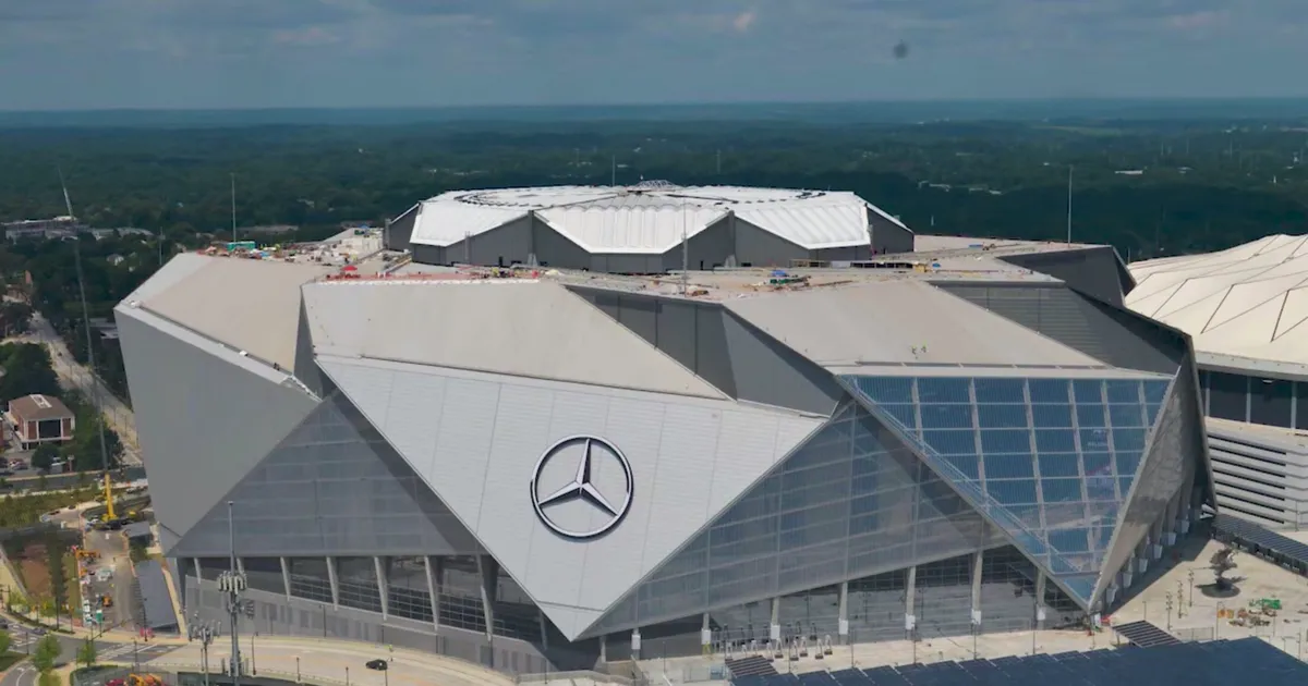 An aerial view of Mercedes-Benz Stadium in Atlanta, its angular silver roof of triangular panels meeting at a circular opening above the field. A large Mercedes-Benz star sits on the sloping glass facade, with green woodland stretching to the horizon behind the stadium.