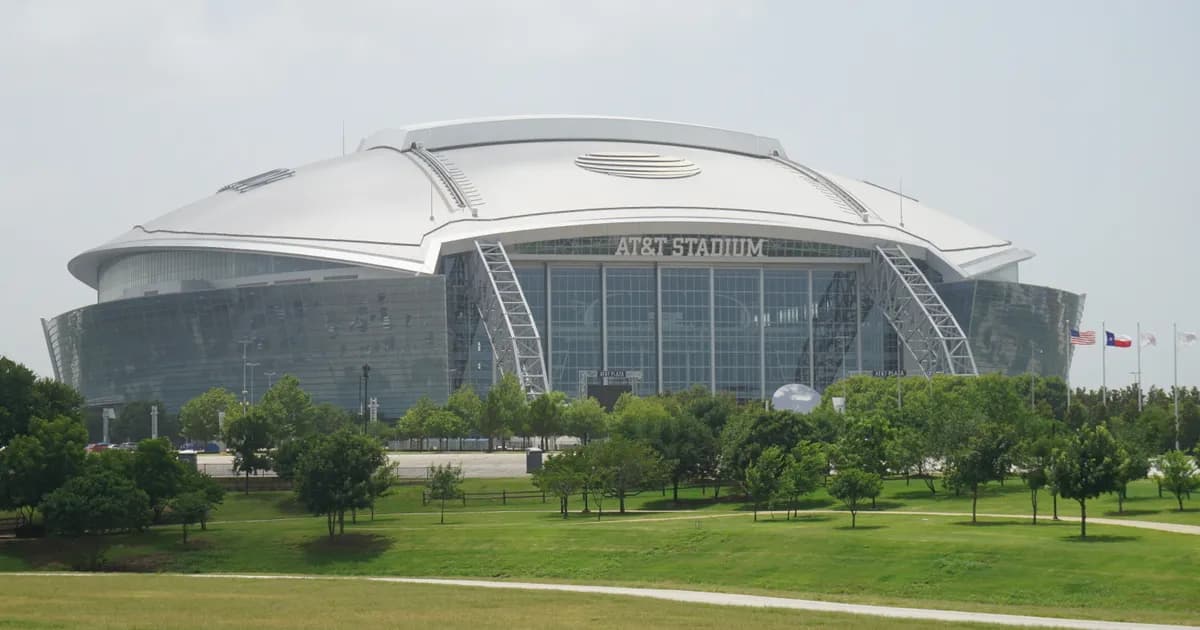 AT&T Stadium in Arlington, Texas A ground-level view of AT&T Stadium in Arlington, Texas, a vast domed stadium with a curved silver roof and dark glass walls, its name across the front. Green lawn and trees sit in the foreground under a pale sky.