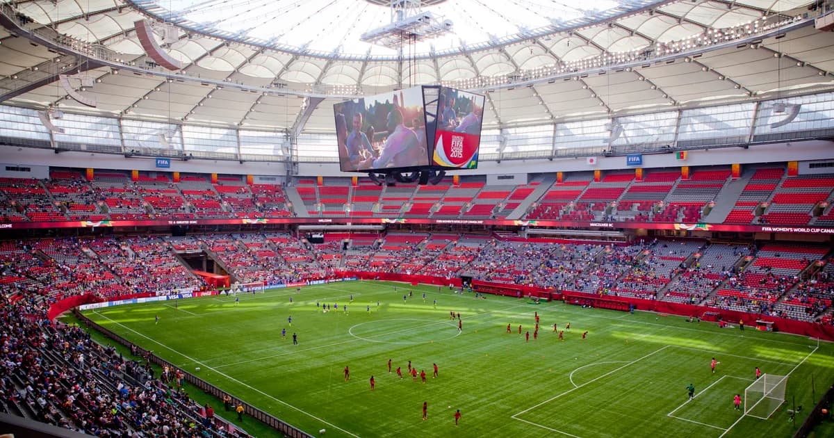 BC Place stadium in Vancouver A view from the upper stands of BC Place in Vancouver during a soccer match, the green pitch below ringed by red seats under a vast white domed roof. A large scoreboard hangs from the center of the stadium roof.