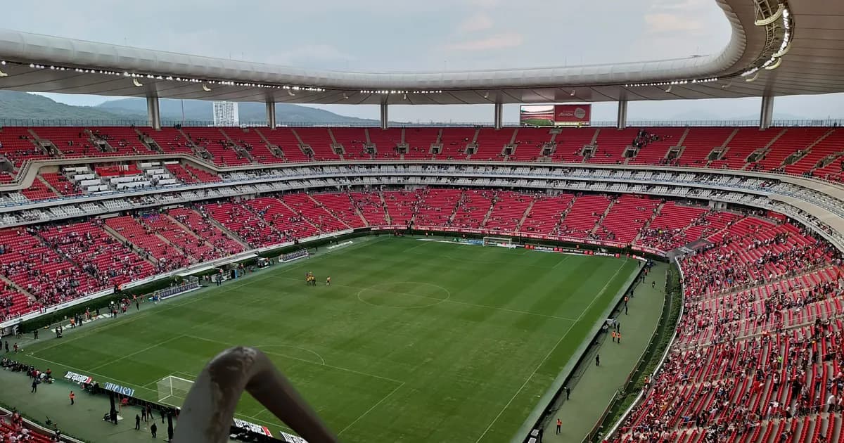 Estadio Akron near Guadalajara A view from the stands of Estadio Akron in Zapopan, near Guadalajara, its tiers of red and white seats curving around a green pitch beneath a wide white roof. Green hills rise beyond the open ends of the stadium.