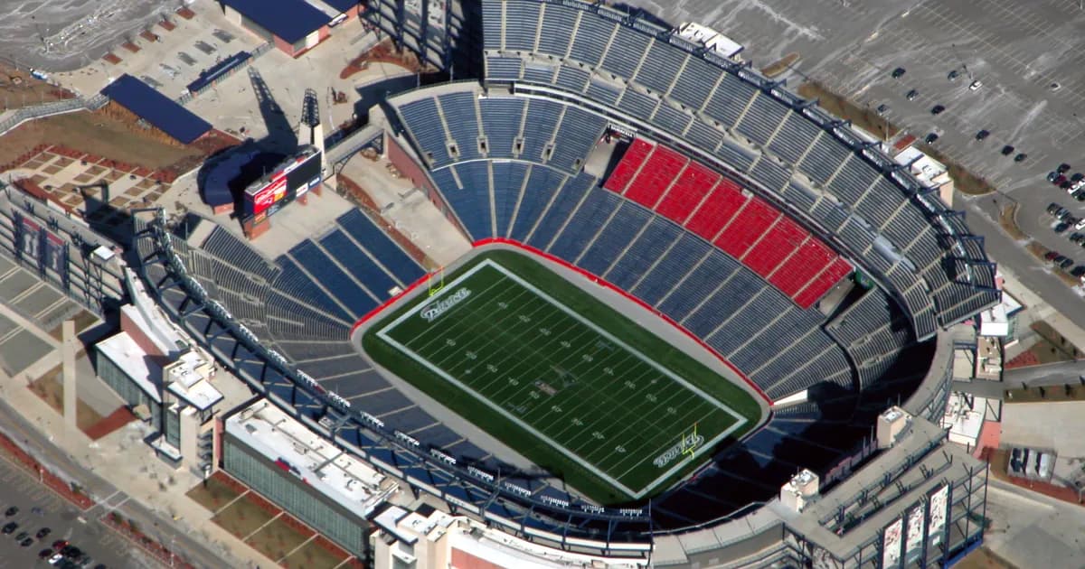 Gillette Stadium in Foxborough, Massachusetts An aerial view of Gillette Stadium in Foxborough, Massachusetts, its grey seating bowl with a block of red seats wrapping a green field. A lighthouse-style tower and wide parking lots sit beside the stadium.