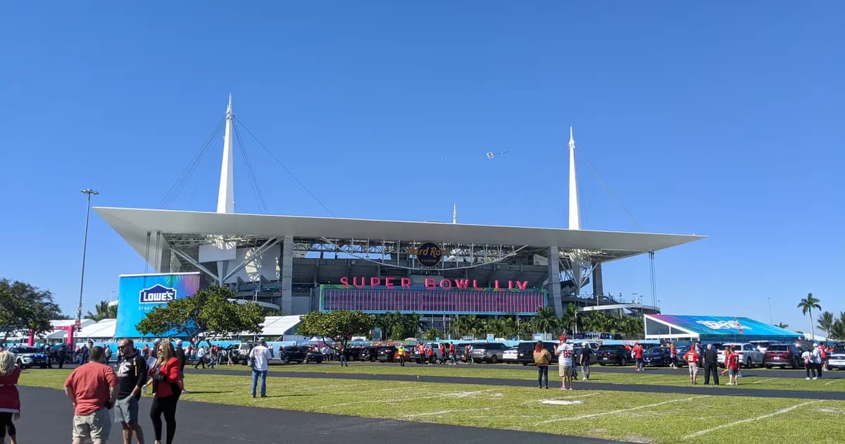 Hard Rock Stadium in Miami Gardens, Florida A view of Hard Rock Stadium in Miami Gardens, Florida, its broad white canopy roof held up on tall masts above the stands, with event signage on the facade. Fans cross a grassy plaza toward the entrance under a clear blue sky.
