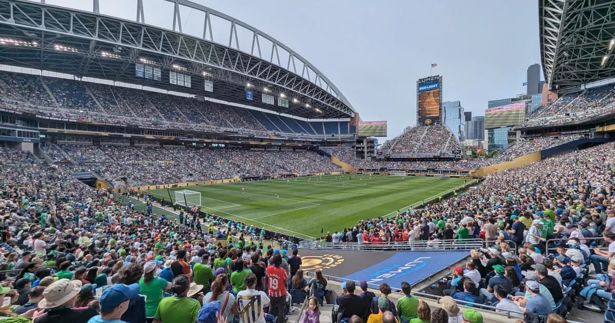 Lumen Field in Seattle A view from the stands of a packed Lumen Field in Seattle during a soccer match, fans filling every tier around the green pitch. The open end of the stadium frames the Seattle skyline under a grey sky.