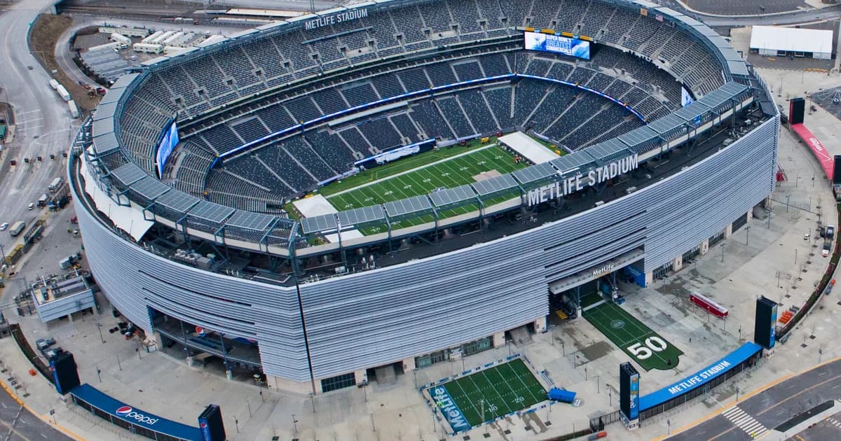 Aerial view of MetLife Stadium near New York An aerial view of MetLife Stadium in East Rutherford, New Jersey, its ribbed silver exterior wrapping an open bowl of dark blue seats around a green football field. The name MetLife Stadium runs along the rim, with highways and parking lots spread around the venue under an overcast sky.