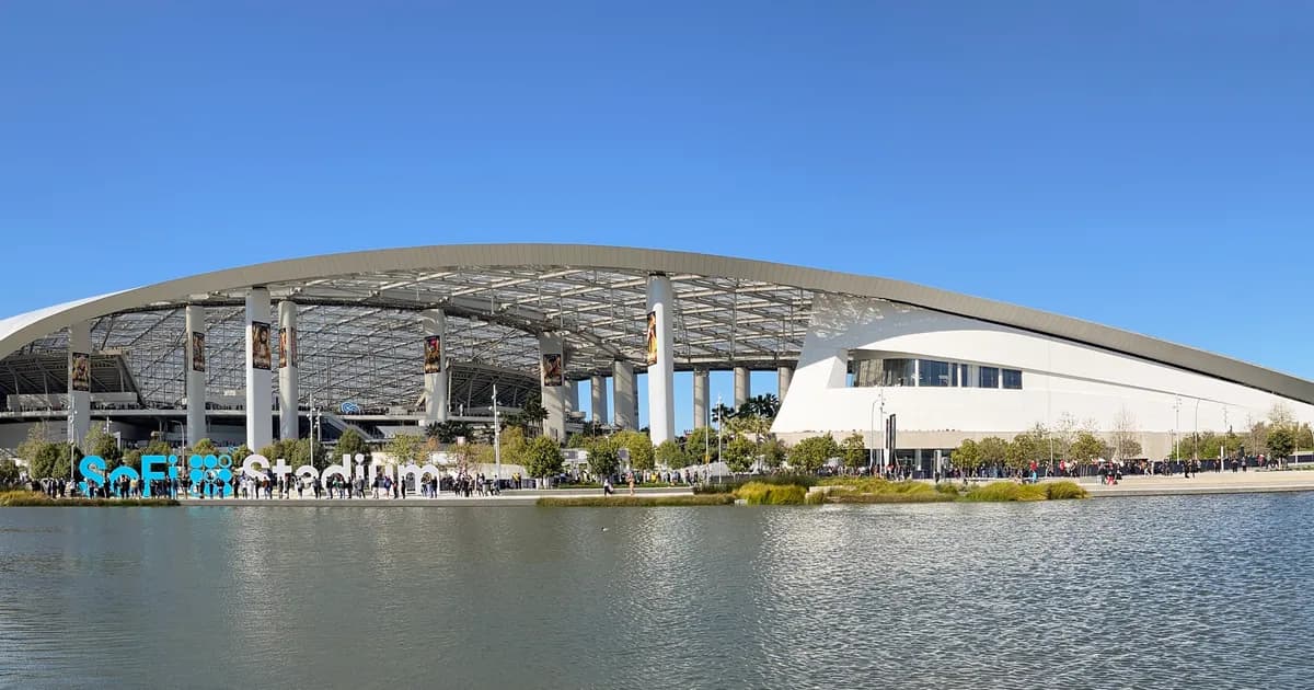 SoFi Stadium in Inglewood, near Los Angeles A wide daytime view of SoFi Stadium in Inglewood, near Los Angeles, its curving translucent-canopy roof spanning an open-sided seating bowl. Crowds walk a landscaped plaza beside a lake, with teal SoFi Stadium letters on the far bank under a clear blue sky.